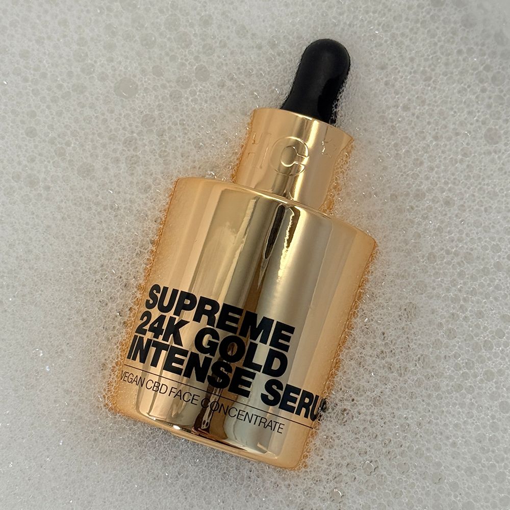 Goldene Serumflasche in Seifenblasen. Aufschrift: Supreme 24K Gold Intense Serum. Schwarze Pipette.