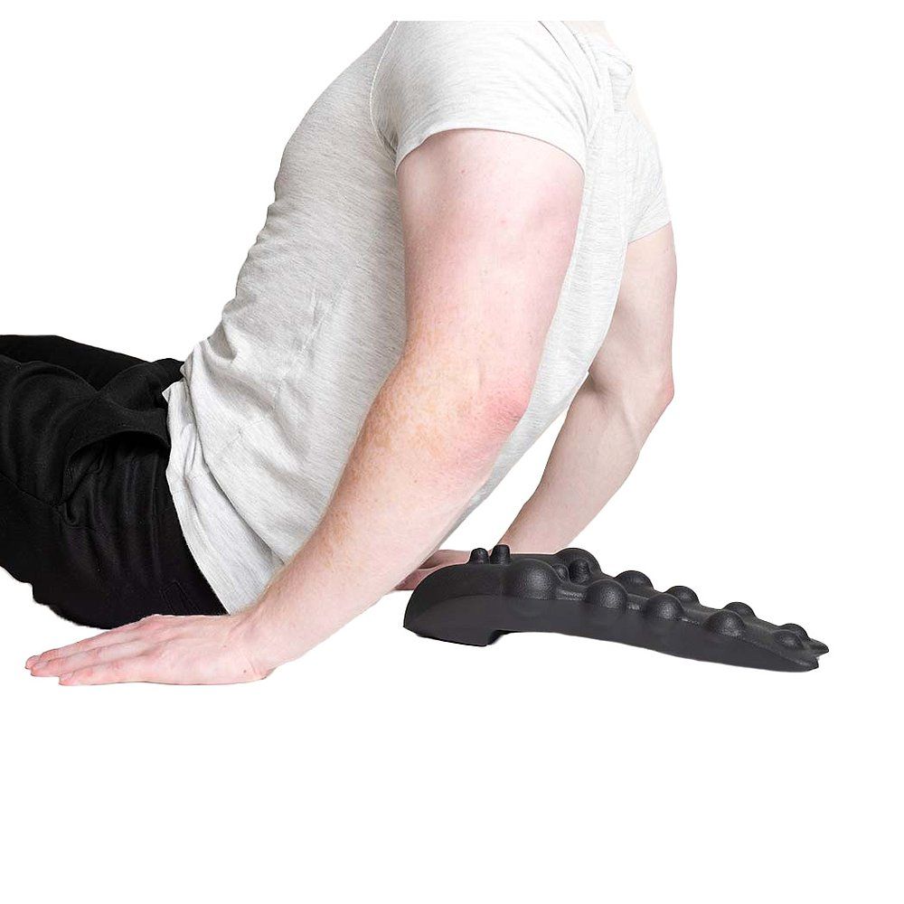 Person sitzt. Ein schwarzes Massagegerät mit Noppen liegt neben der Person. Person trägt ein weißes T-Shirt.