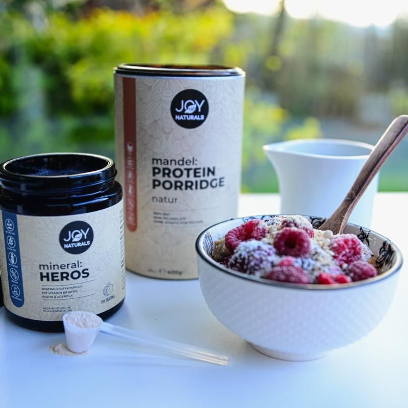 Zwei Dosen und eine Schüssel mit Porridge und Beeren. Eine Dose mit "JOY NATURALS mineral: HEROS", die andere mit "mandel: Protein Porridge".