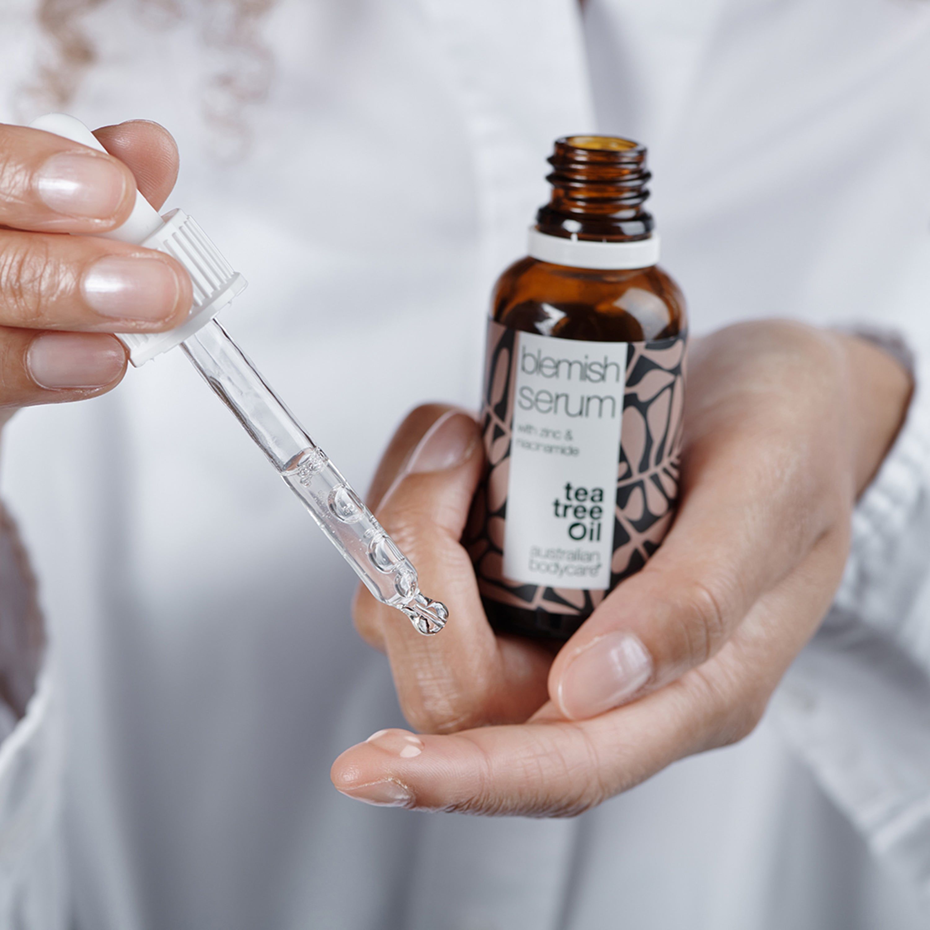 Hände halten Serumflasche und Pipette. Aufschrift: Blemish Serum, Tea Tree Oil, Australian Bodycare.
