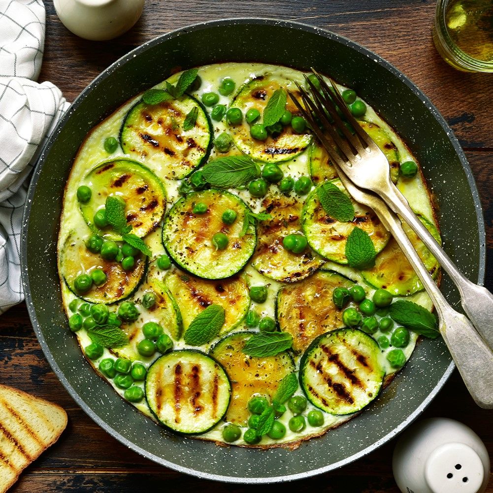 Omelett in einer Pfanne mit gegrillten Zucchinischeiben, Erbsen und Minze. Zwei Gabeln liegen daneben.