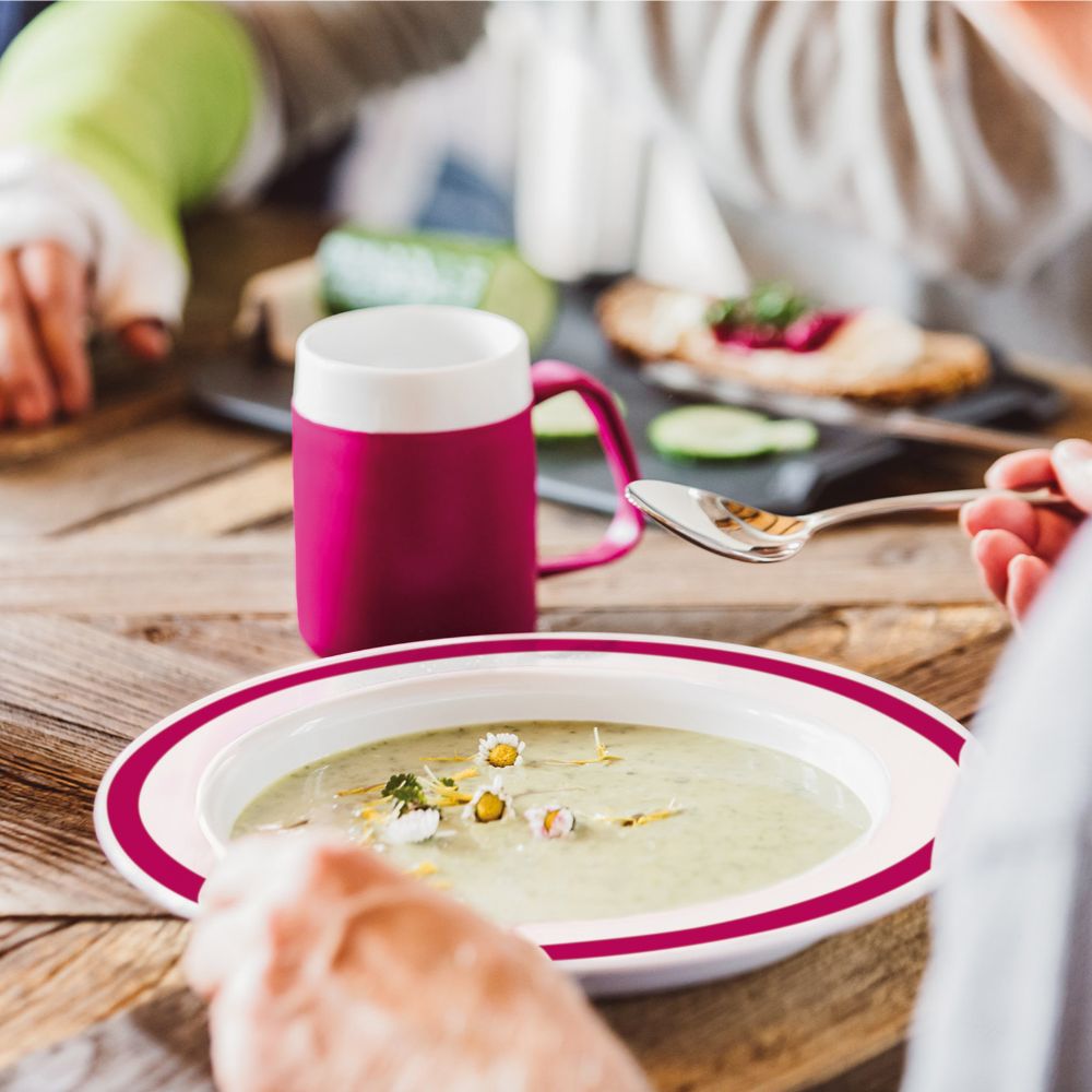 Teller mit Suppe, weiß mit pinkfarbenem Rand. Löffel wird in die Suppe getaucht. Auf Holztisch. Person hält Tasse.