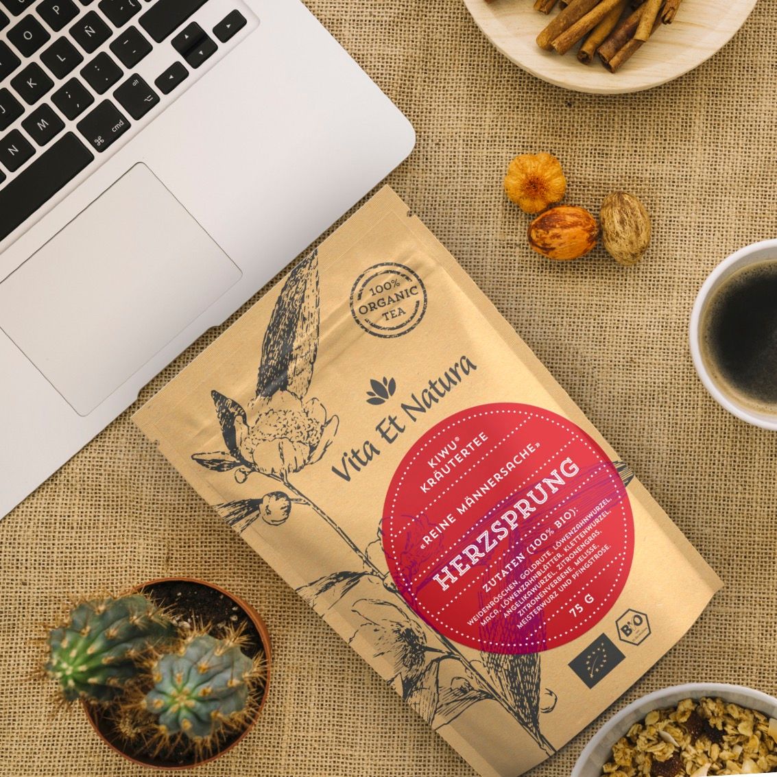 Teeverpackung auf Tisch mit Laptop, Tasse Kaffee und Dekoration. Auf dem Beutel steht: Vita Et Natura, Kräutertee Herzsprung, 100% Bio.