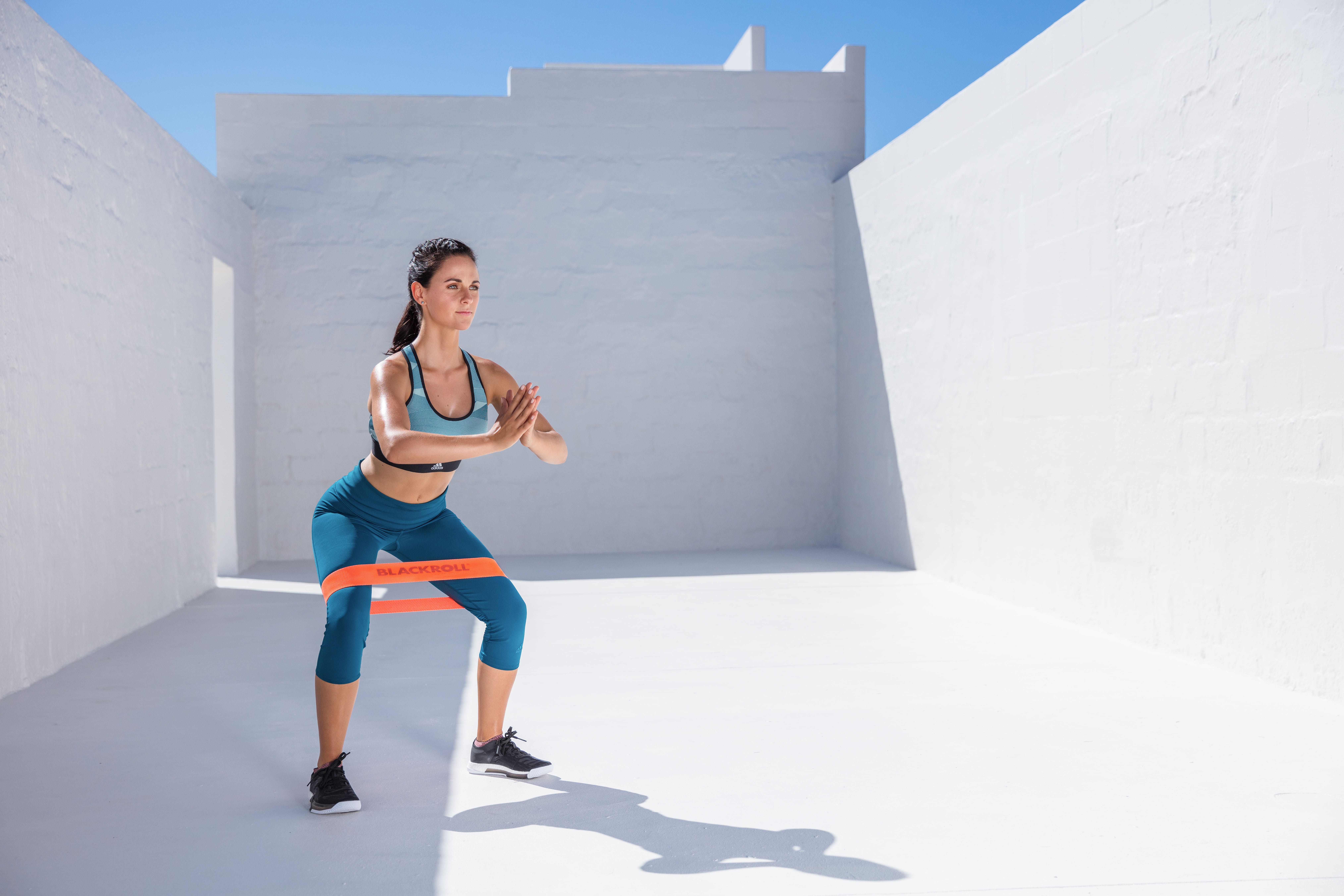 Frau in Sportkleidung macht Kniebeugen mit orangefarbenem Loop Band um die Beine.