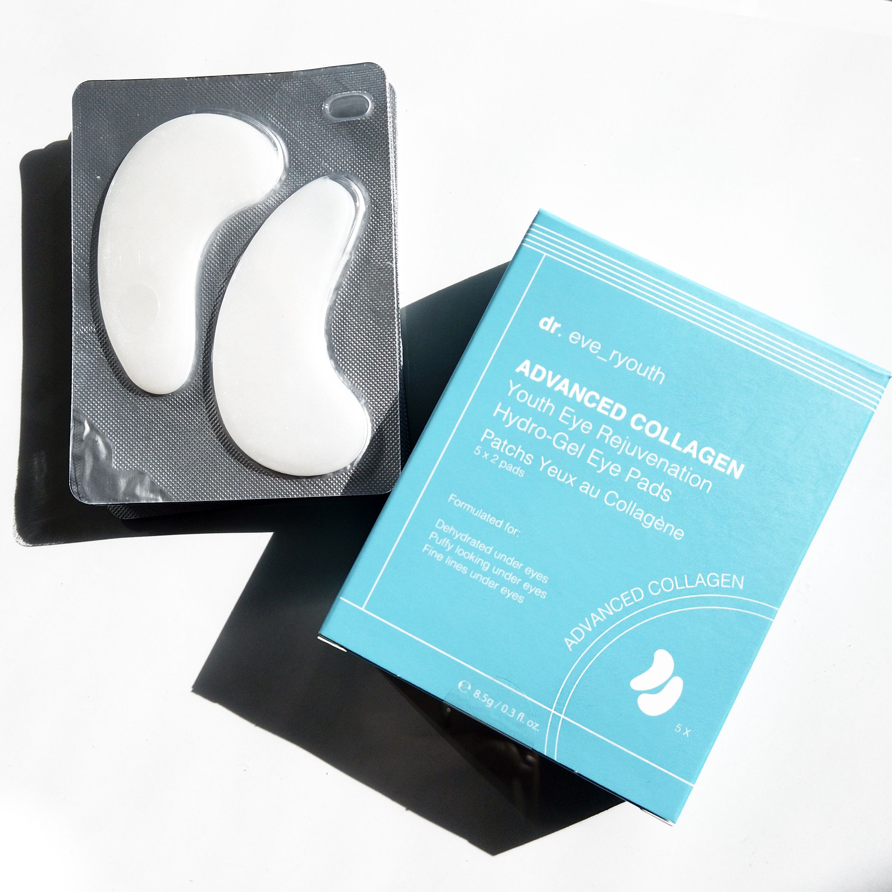 Augenpads in Verpackung und Schachtel. Weiße, halbmondförmige Pads. Blaue Schachtel mit Produktinformationen.