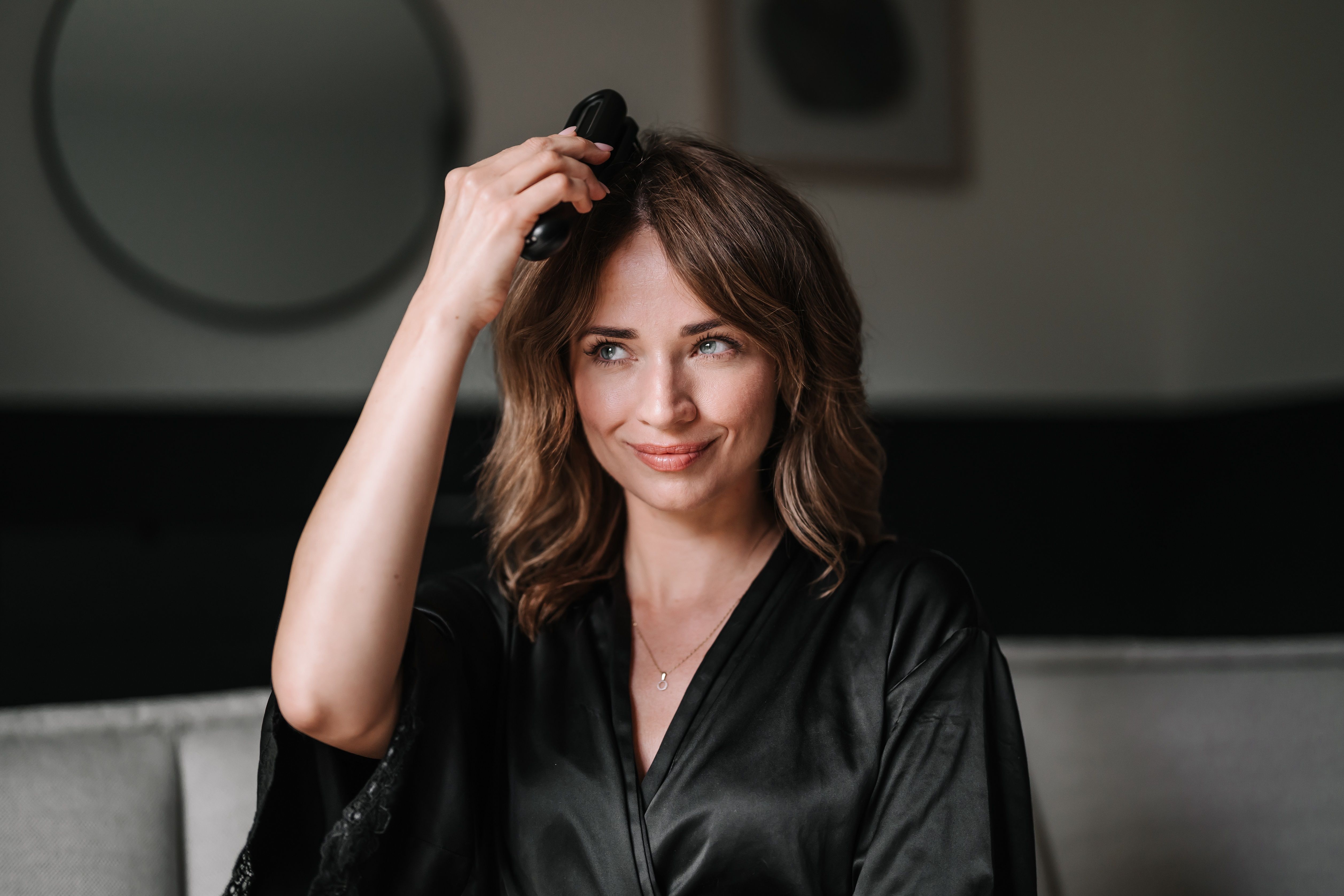Frau mit schwarzem Morgenmantel hält ein schwarzes Beauty-Gerät. Blick in die Kamera. Hintergrund: Wohnzimmer.