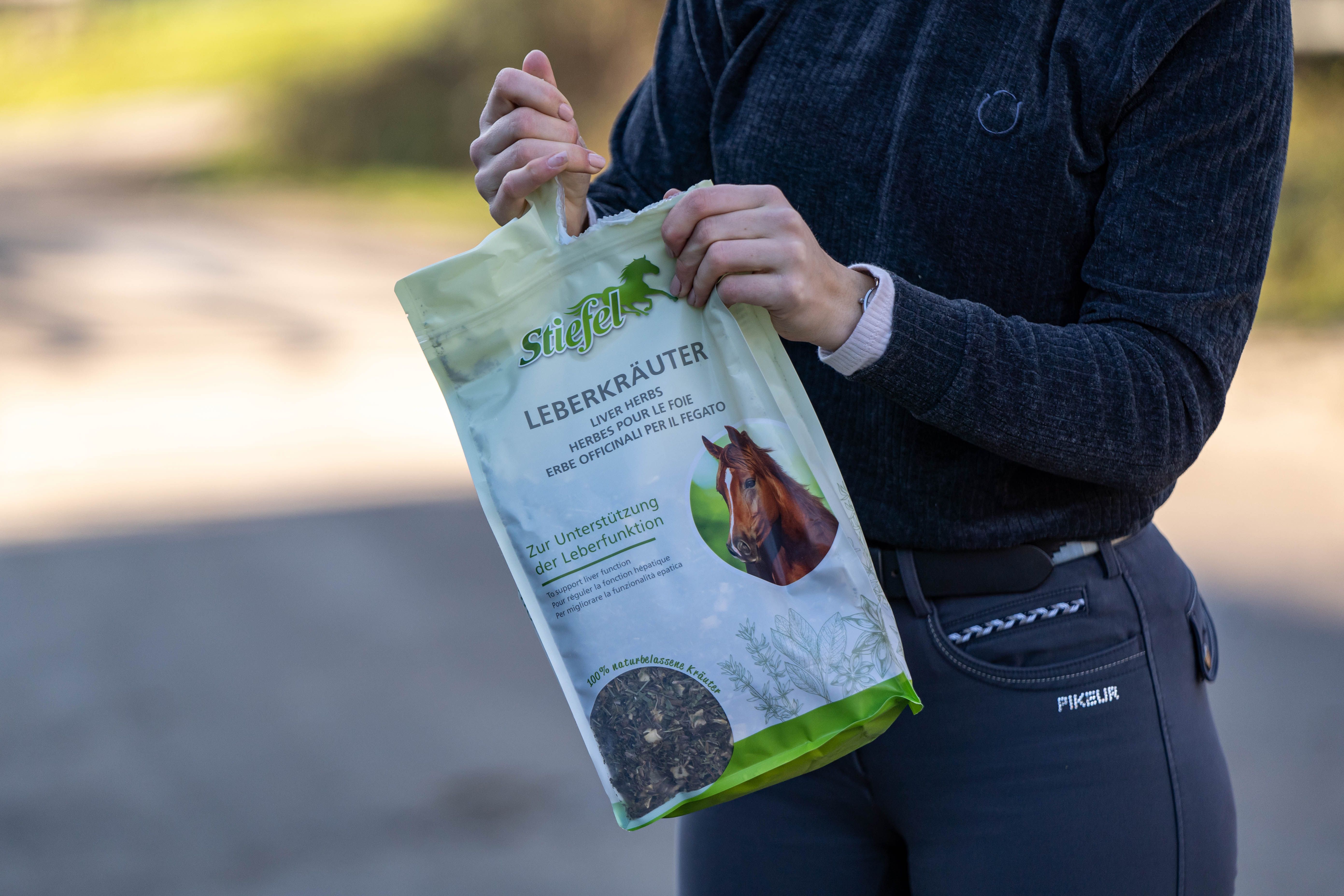 Person hält einen Beutel Stiefel Leberkräuter. Aufdruck: Kräutermischung für die Leberfunktion. Grün-weiße Verpackung, Pferd-Illustration.