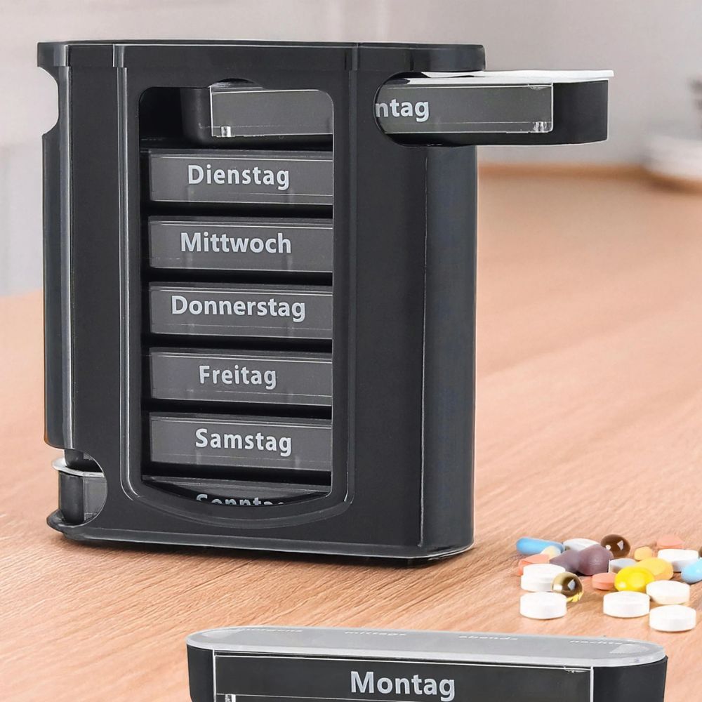 Schwarze Tablettenbox mit 7 Tagesfächern. Ein Fach mit der Aufschrift Montag ist herausgenommen. Tabletten liegen auf dem Tisch.
