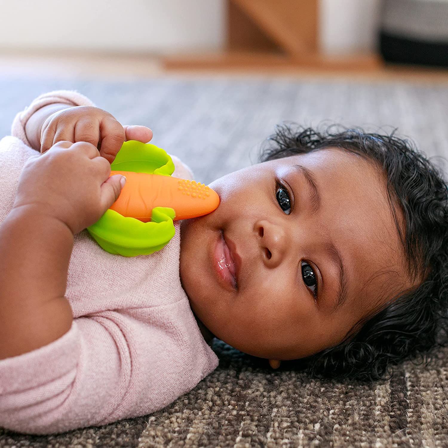 Baby liegt auf dem Boden und hält einen orangefarbenen und grünen Karotten-Beißring. Nahaufnahme des Gesichts.