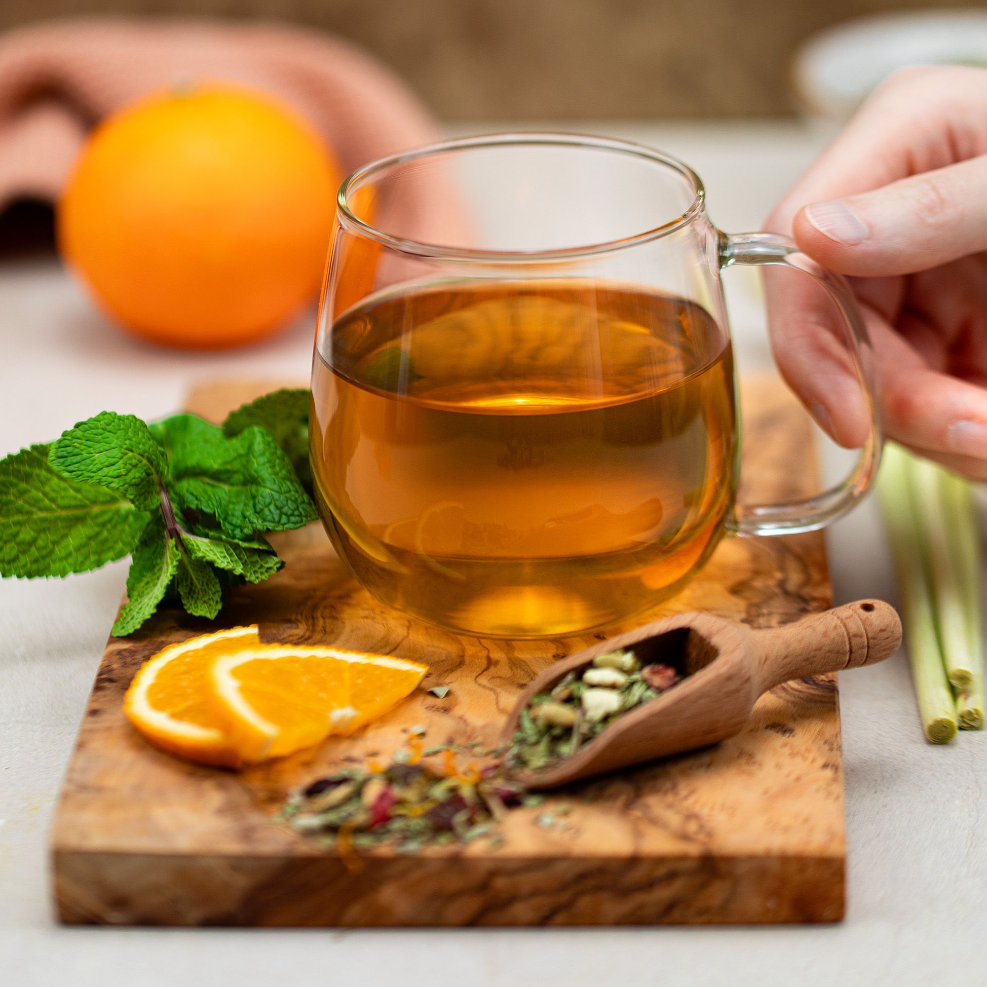 Glasbecher mit Tee auf Holzbrett. Hand greift nach Becher. Holzlöffel mit Kräutern, Orangenscheiben und Minze. Helles Ambiente.
