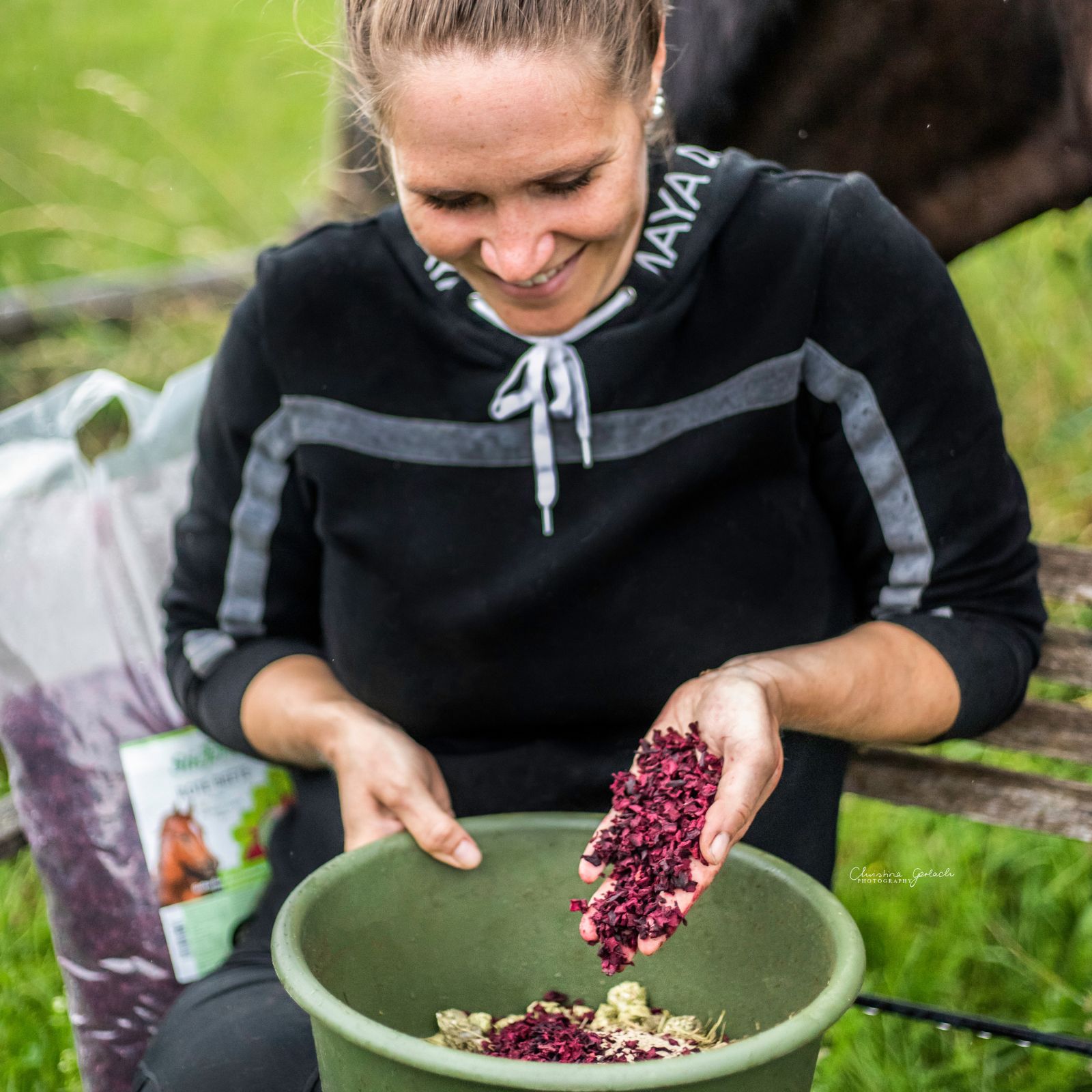 Frau hält eine Handvoll Rote Beete Flocken über einer grünen Schüssel. Im Hintergrund eine weitere Packung und ein Pferd.