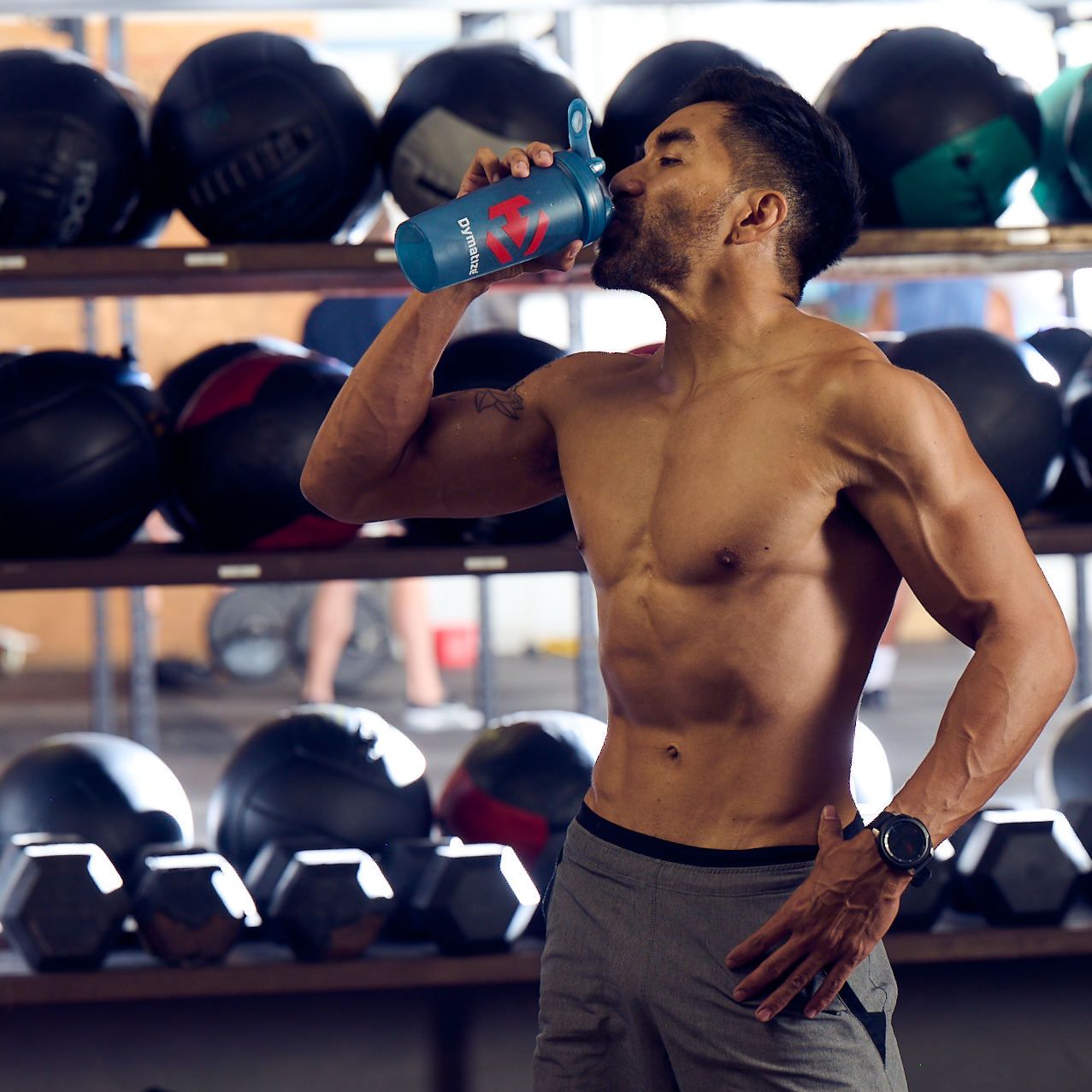 Ein Mann trinkt aus einer blauen Shaker-Flasche in einem Fitnessstudio mit Gewichten im Hintergrund.