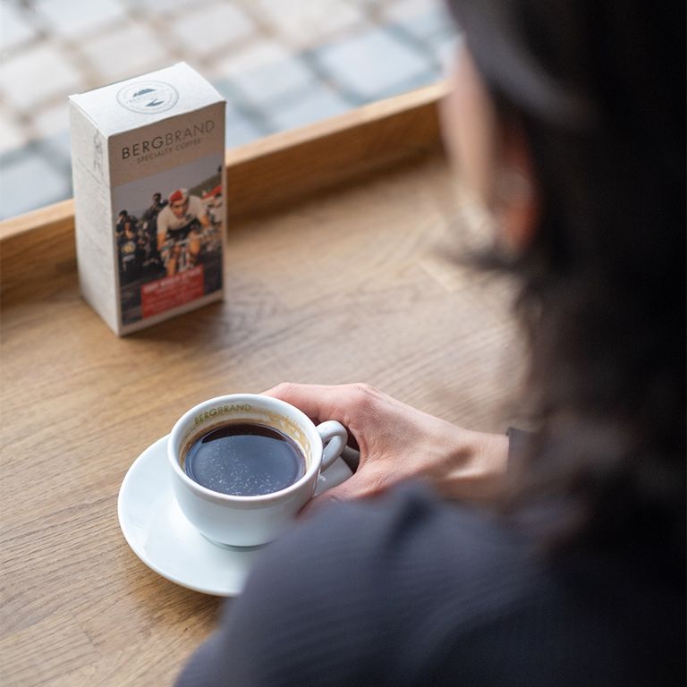 Kaffeetasse mit Kaffee und Kaffeepackung BERGBRAND Eddy would Attack. Person im Hintergrund. Auf einem Tisch.