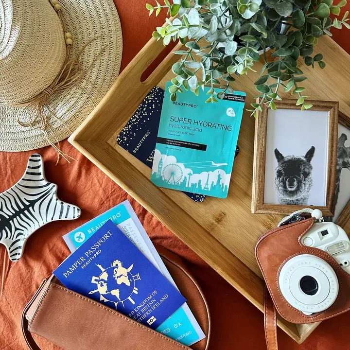 Turquoise Beautypro-doekmasker op een dienblad met reisaccessoires. Opschrift: Super Hydrating Hyaluronic Acid. Verpakking met stadsgezicht.