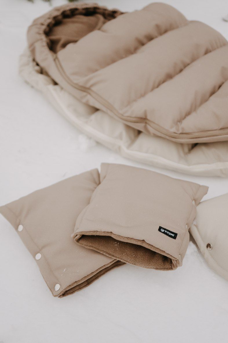 Ein wattierter, beiger Babyschlafsack und passende Fäustlinge liegen im Schnee. Ein Etikett mit dem Schriftzug 'TTOMI' ist auf einem Fäustling sichtbar.
