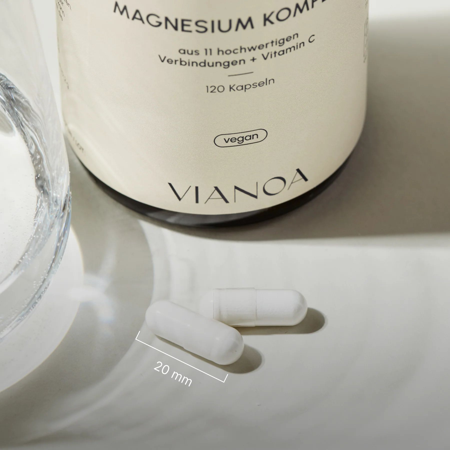 Zwei VIANOA Magnesium Komplex Kapseln (20 mm) neben einem Glas Wasser und der Produktflasche.
