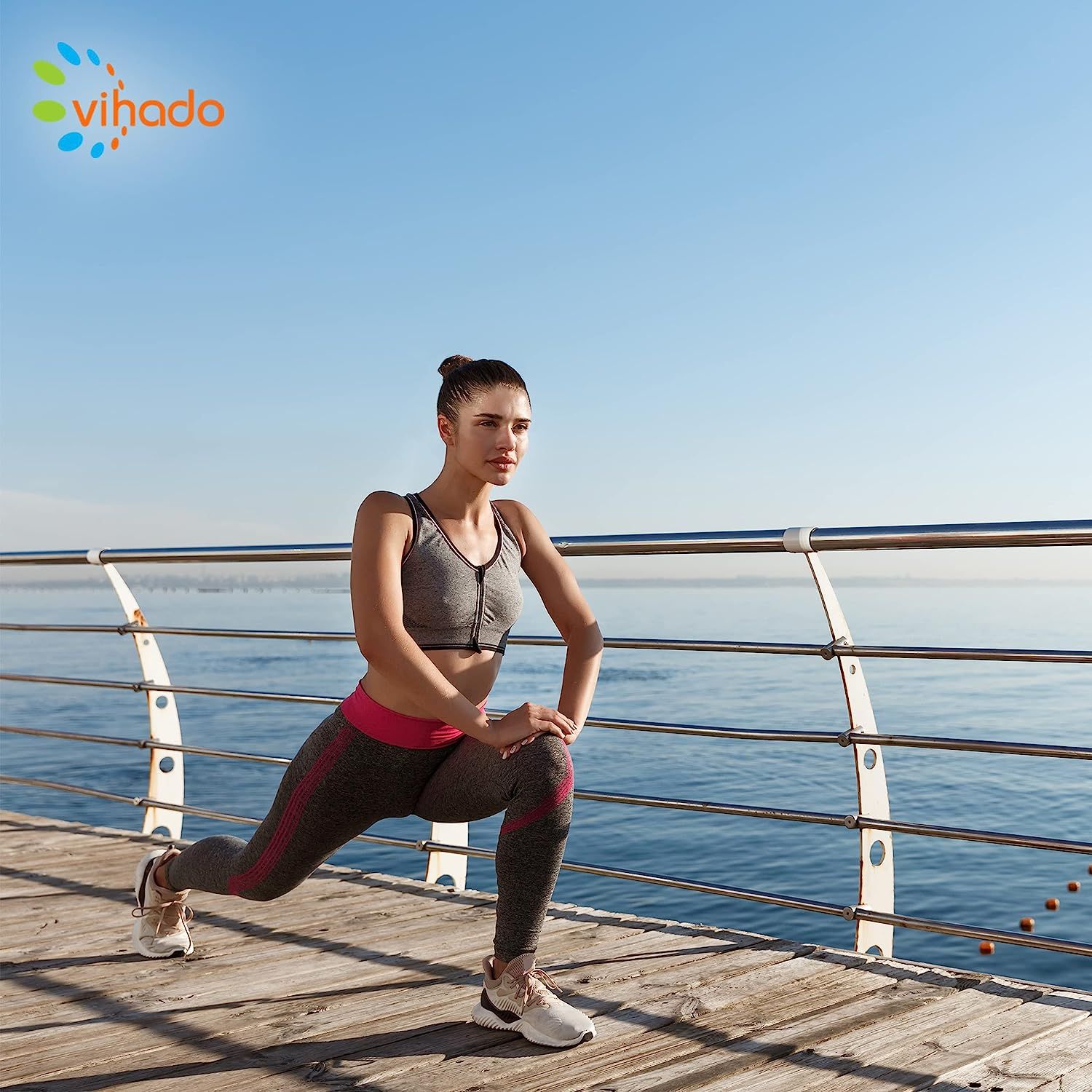 Frau in Sportkleidung dehnt sich auf einem Steg am Meer. Graues Top, Leggings. Hintergrund: Meer und blauer Himmel.