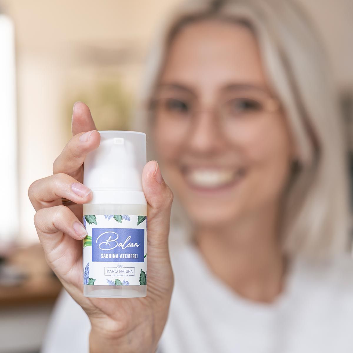 Person hält Balsam-Spender. Aufschrift: Balsam Sabrina Atemfrei. Karo Natura Logo. Person trägt Brille und lächelt.