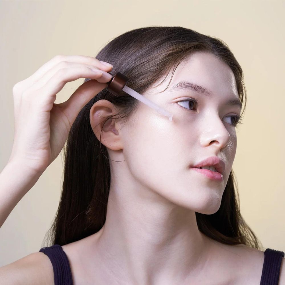 Frau hält Pipette mit Serum. Gesicht, Arme und Schultern sind sichtbar. Hintergrund ist hellgelb.