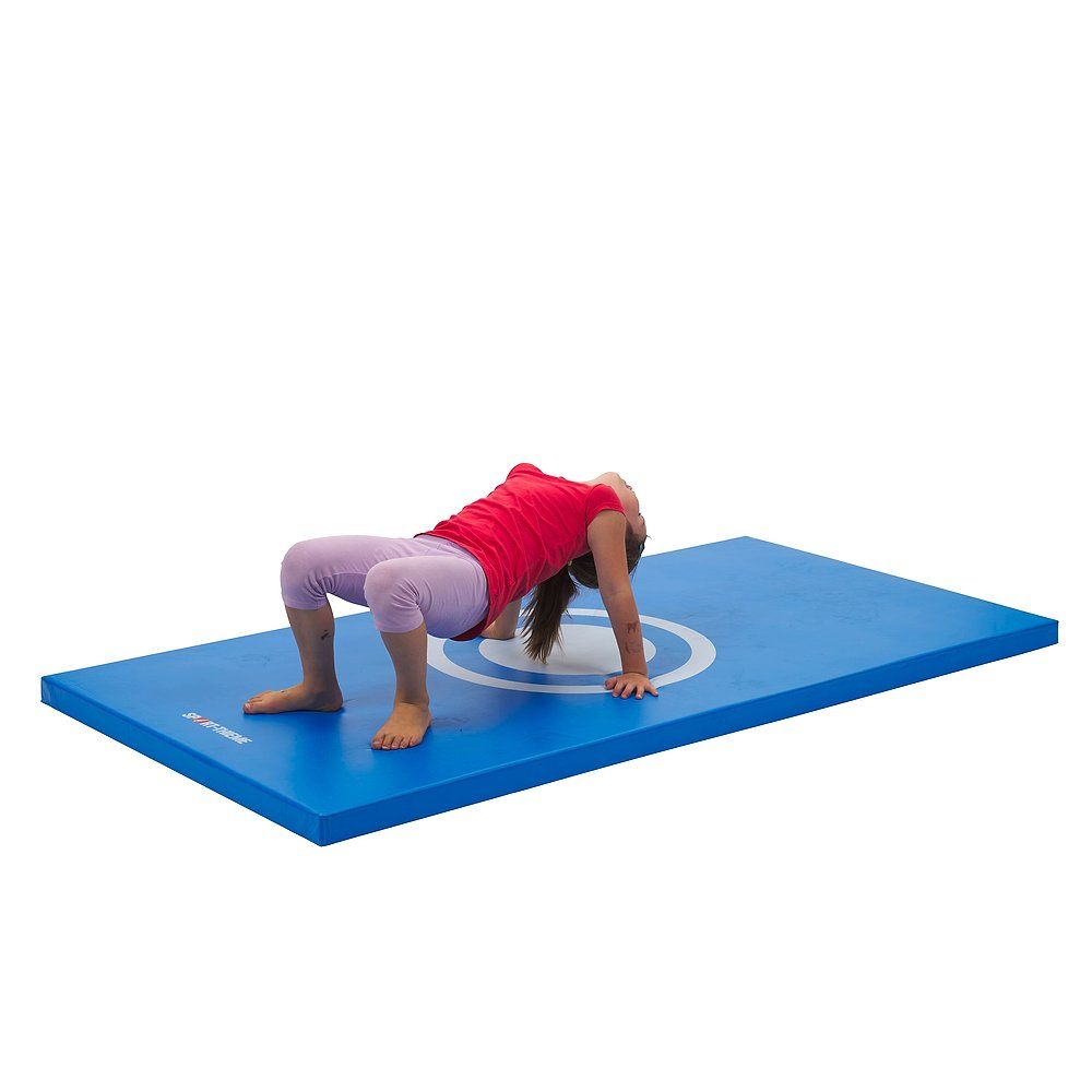 Blaue Turnmatte mit weißem Zielkreis. Ein Mädchen macht eine Brücke auf der Matte. Sie trägt ein rotes Oberteil und eine rosa Hose.