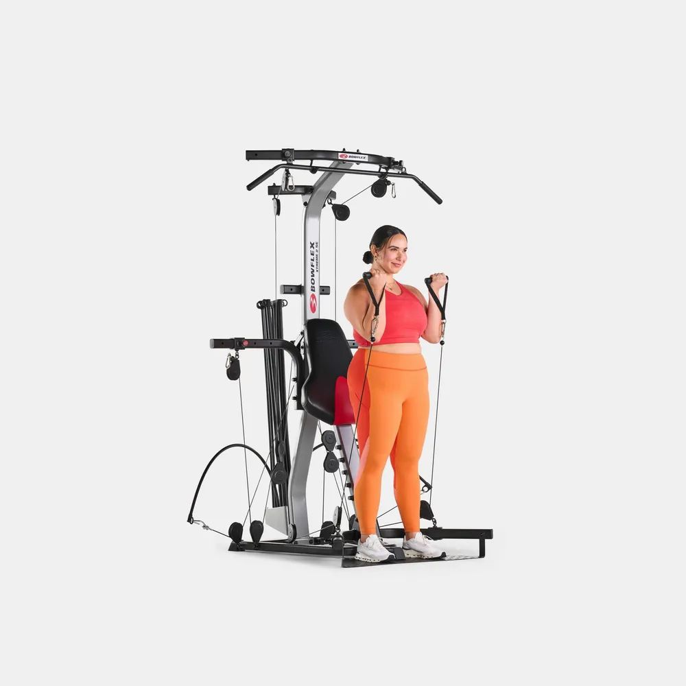 Frau trainiert an einem Fitnessgerät. Rotes Oberteil, orangefarbene Hose. Graues Fitnessgerät mit schwarzen Elementen.