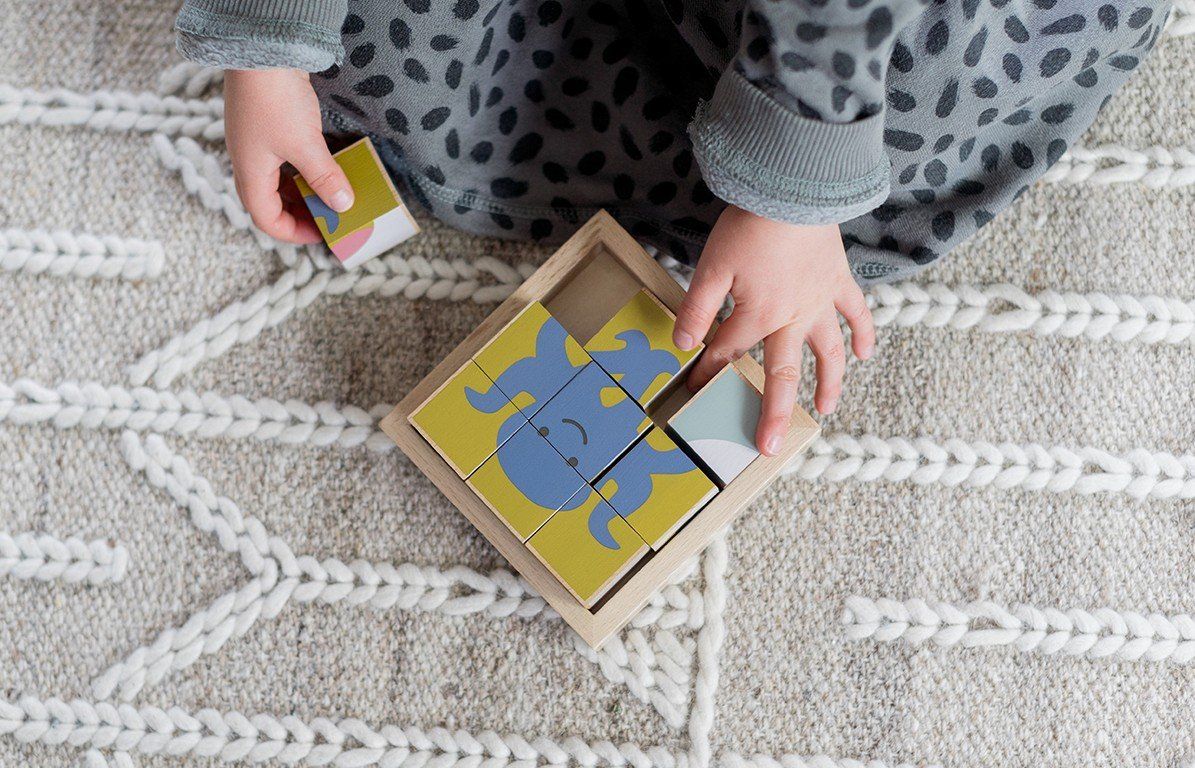 Holzwürfel-Puzzle mit Elefanten-Motiv. Neun Würfel bilden ein Bild in einem Holzrahmen. Gelb, blau und grau. Kind hält Würfel.