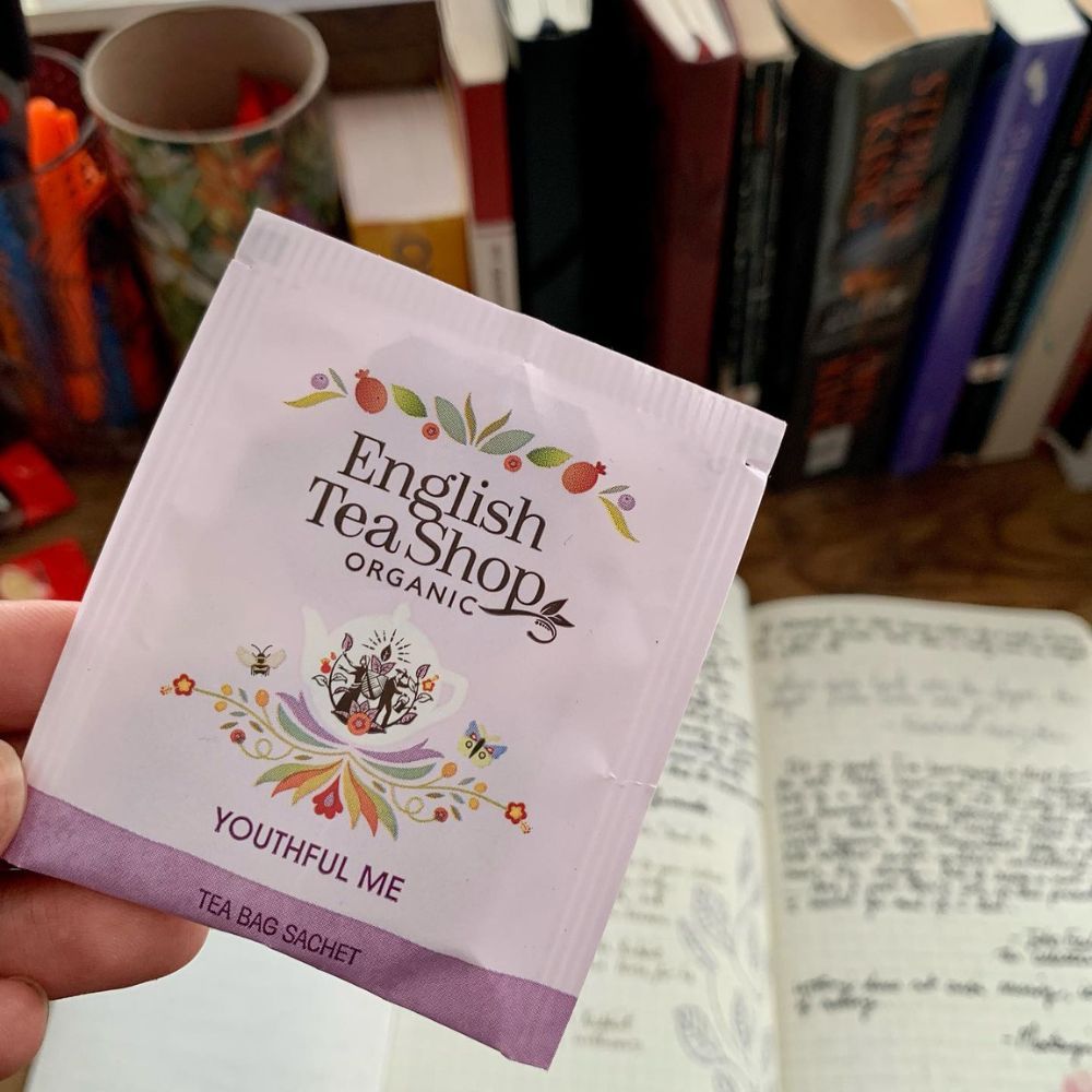 Einzelner Teebeutel ETS - Youthful Me. Aufdruck: English Tea Shop Organic. Beutel wird gehalten. Hintergrund: Notizbuch, Bücher.