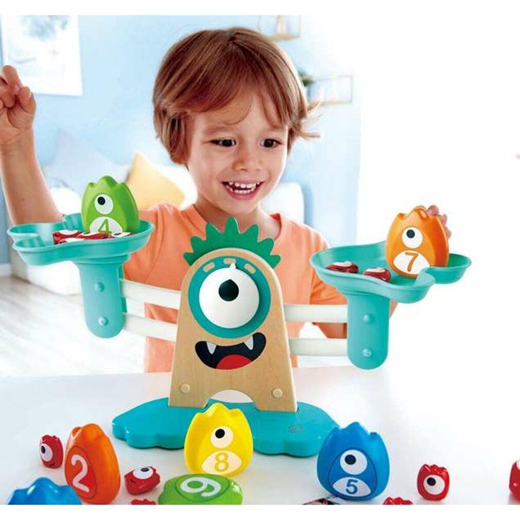 Ein Kind spielt mit der Hape Monster-Waage. Die Waage hat eine Holzbasis und blaue Waagschalen. Monster-Gewichte und Zahlen liegen daneben.