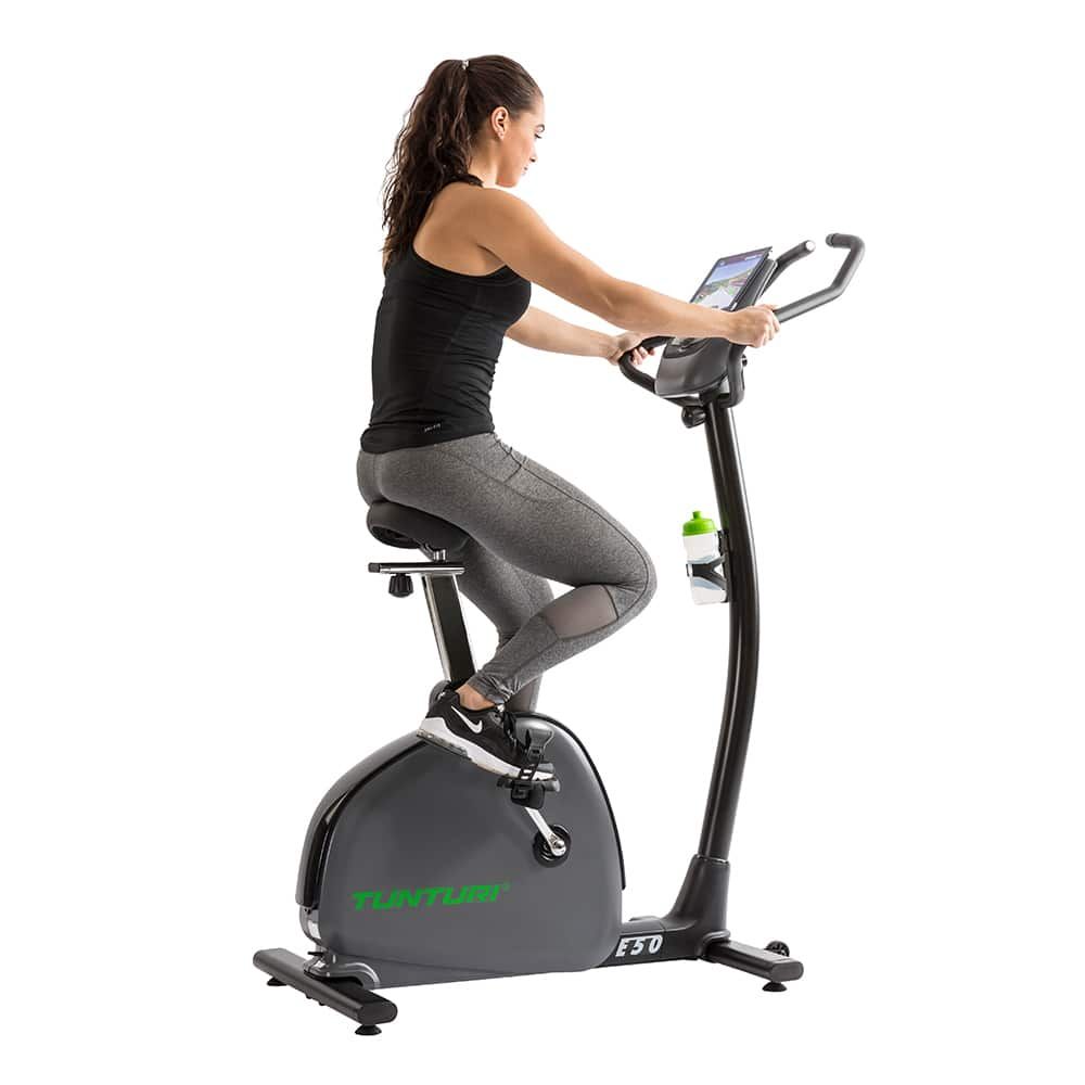 Frau trainiert auf Heimtrainer E50 Performance Bike. Sie sitzt auf dem Sattel, Hände am Lenker. Flasche im Halter.
