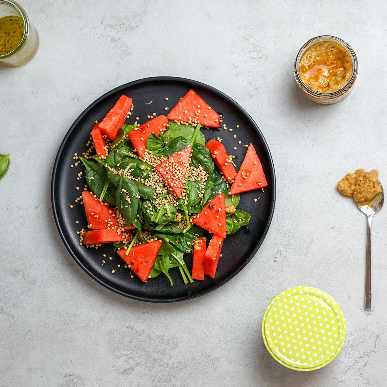 Hummus-Glas neben Salat mit Wassermelone. Aufschrift nicht lesbar.