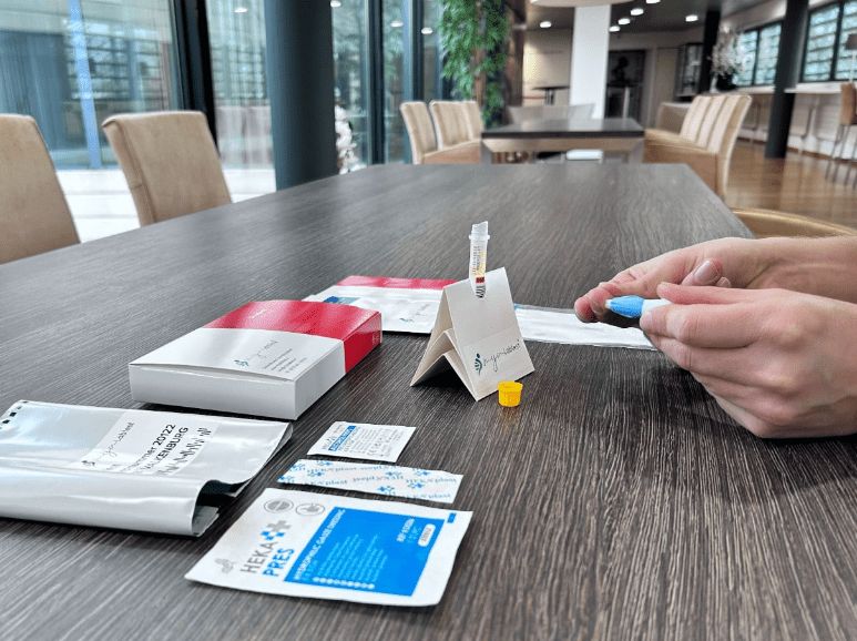 Person hält Lanzette und Röhrchen. Auf dem Tisch: Test-Kit-Komponenten, Meinlabtest-Logo, Heka Pres-Verpackung, Tischplatte.