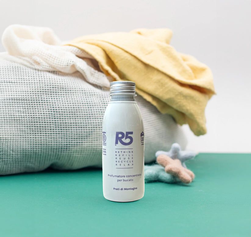 Weißes Fläschchen mit silbernem Deckel. Aufschrift R5 und Text. Produktname unten. Textilwaren im Hintergrund.