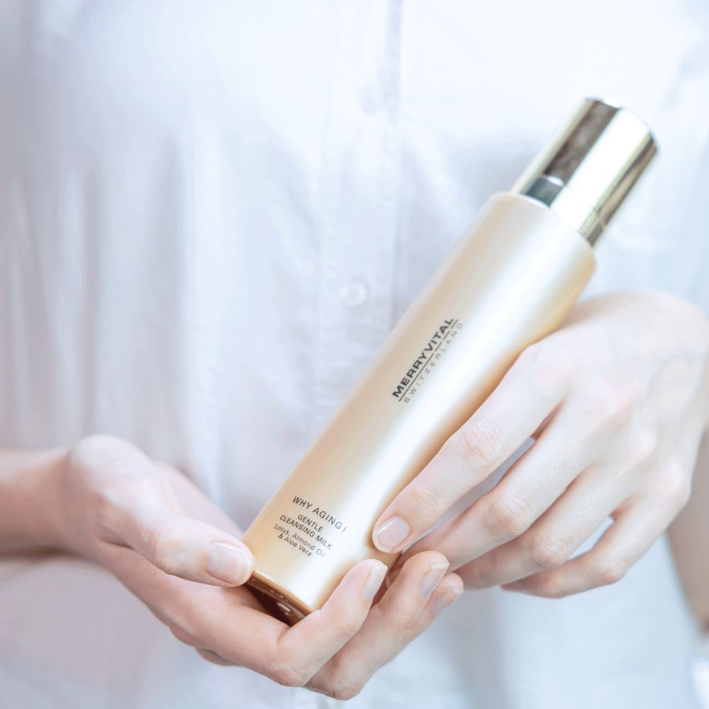 Goldfarbene Flasche in Händen. Aufschrift: MERRYVITAL, WHY AGING!, Gentle Cleansing Milk. Helle Kleidung im Hintergrund.