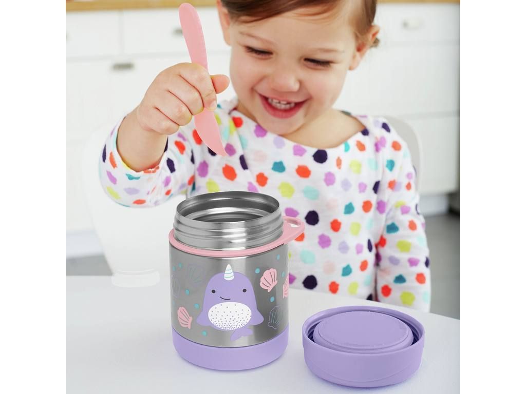Kind hält rosa Löffel über offener Edelstahl-Warmhaltebox mit Narwal-Motiv. Lila Deckel daneben. Tisch mit Essen.