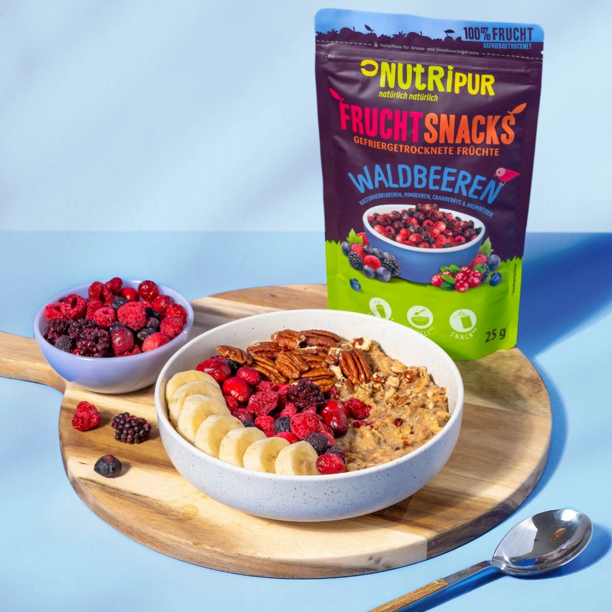 Schüssel mit Müsli, Bananenscheiben, Beeren und Nüssen. Daneben Beutel NutriPur Waldbeeren und Schüssel mit Beeren.