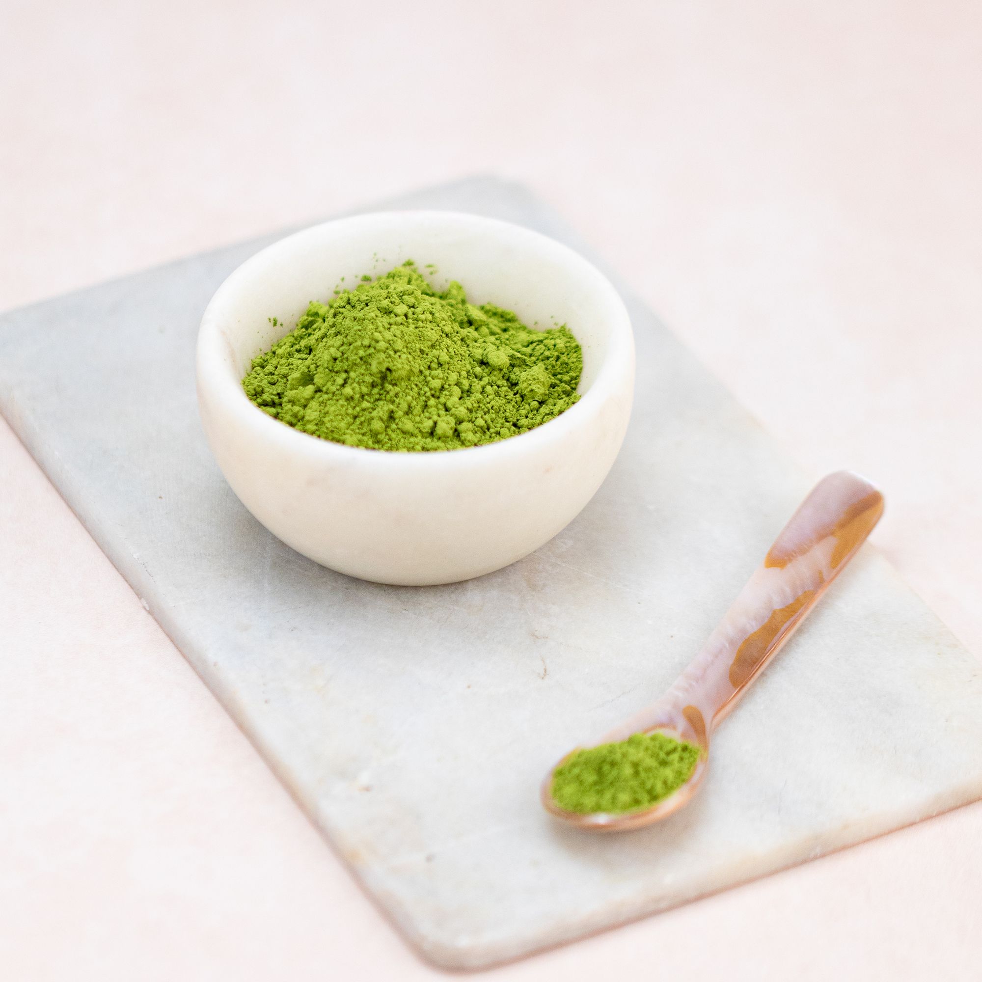 Kleine Schale mit grünem Matcha-Pulver. Löffel mit Matcha-Pulver. Beides auf einer Marmorplatte.