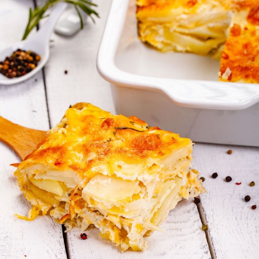 Auflauf auf Holzbrett. Kartoffeln, Käse, Gewürze. Weißer Auflaufform im Hintergrund. Holzbrett mit Auflaufstück.