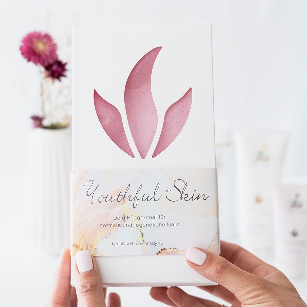 Person hält Kosmetik-Set. Aufschrift: Youthful Skin. Verpackung mit floralem Ausschnitt. Produkte: Super Gel, Secrets Lotion im Hintergrund.