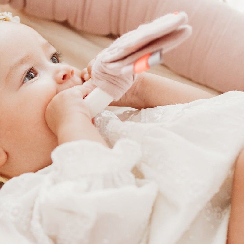 Baby mit Beißring im Mund. Rosa Stoffohren und weißer Ring. Baby trägt weißes Kleid.