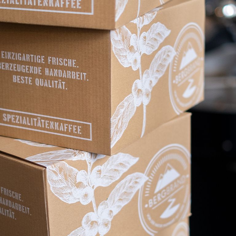 Kaffeeprodukte in Kartons gestapelt. Aufschrift: BERGBRAND, Spezialitätenkaffee. Text: Einzigartige Frische, überzeugende Handarbeit, beste Qualität.