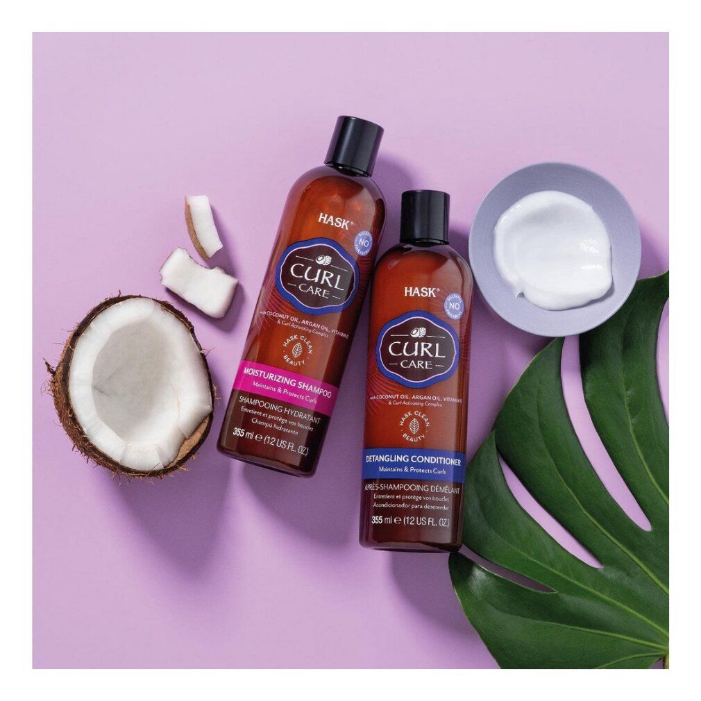 Zwei HASK Curl Care Flaschen, eine mit feuchtigkeitsspendendem Shampoo, die andere mit entwirrendem Conditioner. Kokosnussstücke und eine Schale mit weißer Creme sind daneben platziert.