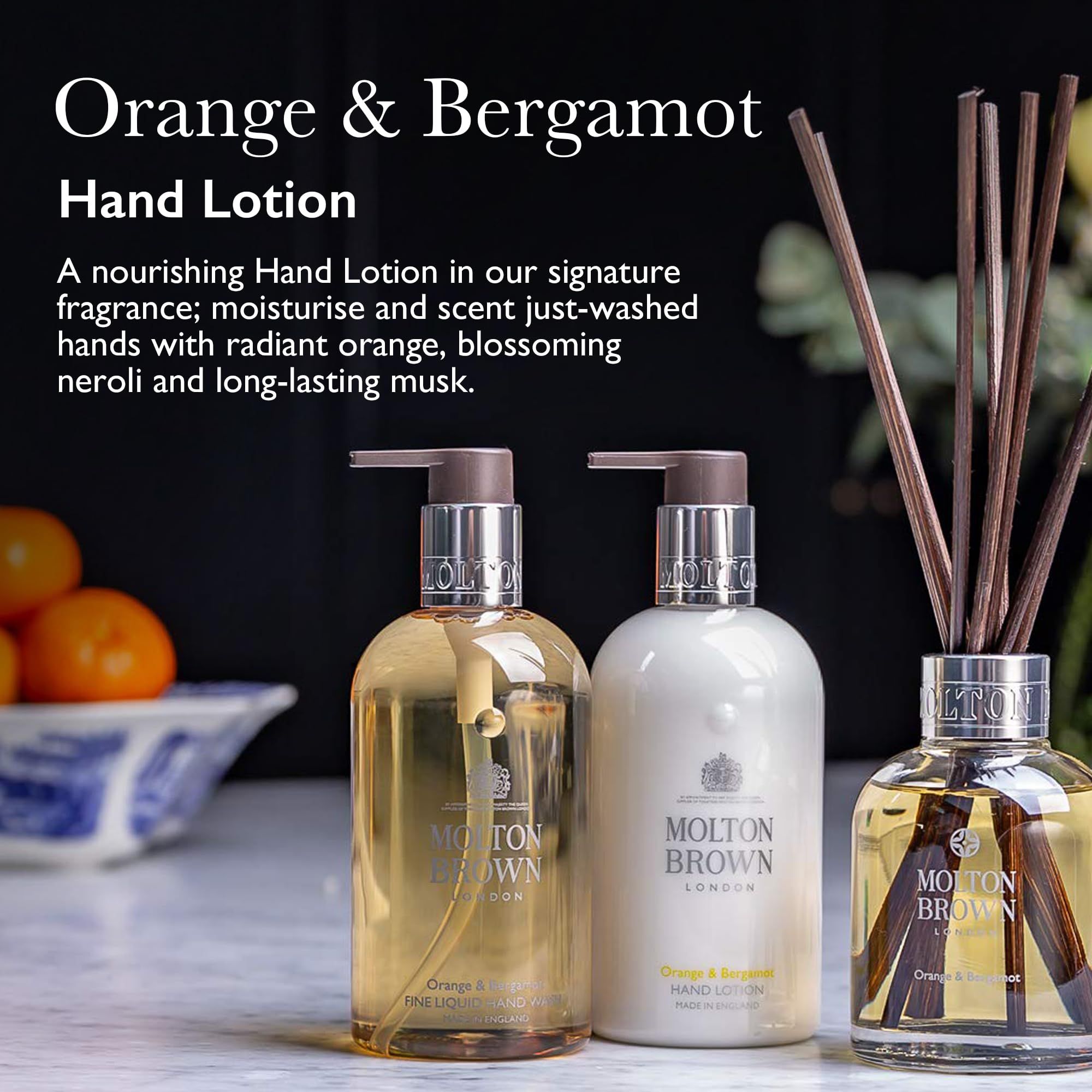 Zwei Flaschen Handlotion und ein Diffusor. Aufschrift: Molton Brown London, Orange & Bergamot. Hintergrund: Obst und Dekoration.