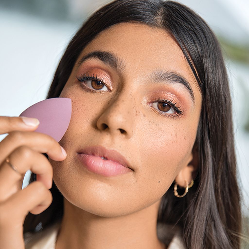 Eine Frau hält einen lilafarbenen Make-up-Schwamm. Der Schwamm ist tropfenförmig. Die Frau berührt ihr Gesicht.