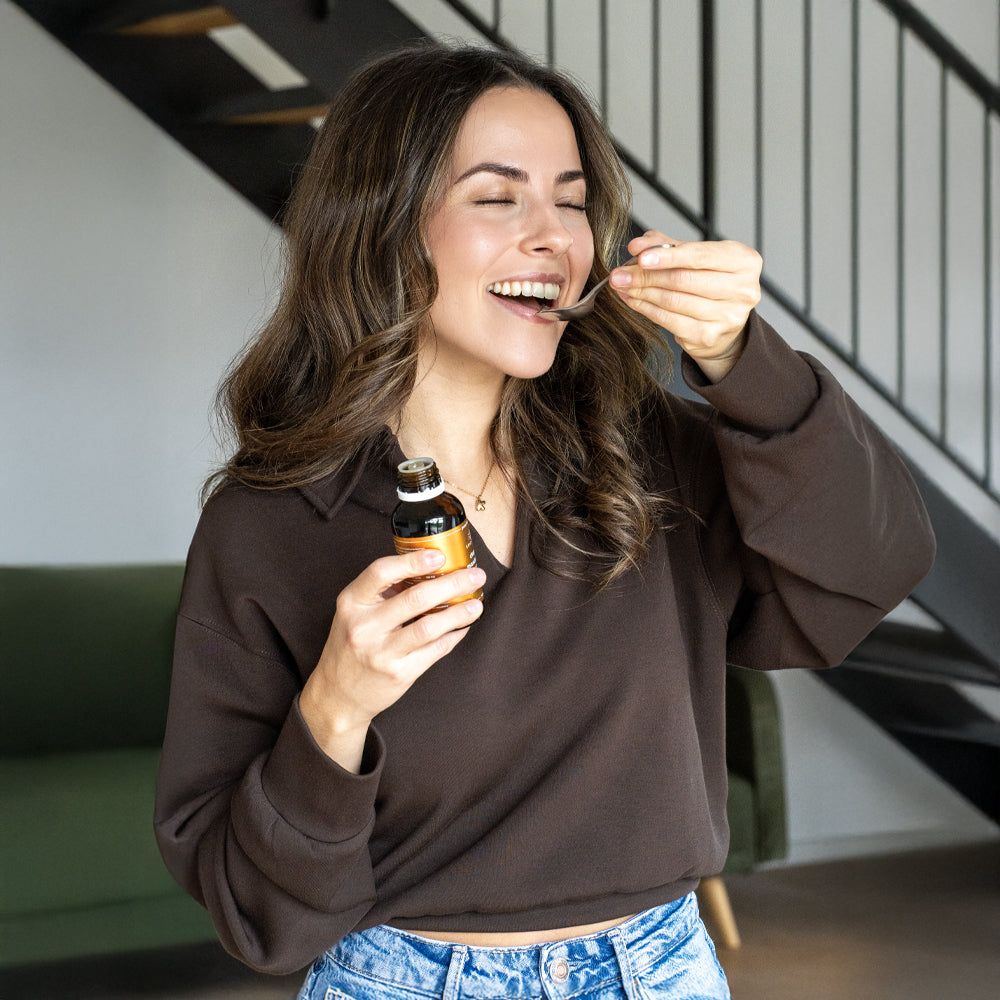 Frau trinkt aus einem Löffel. Flasche mit Produktetikett im Hintergrund. Umgebung: Treppe, Sofa.
