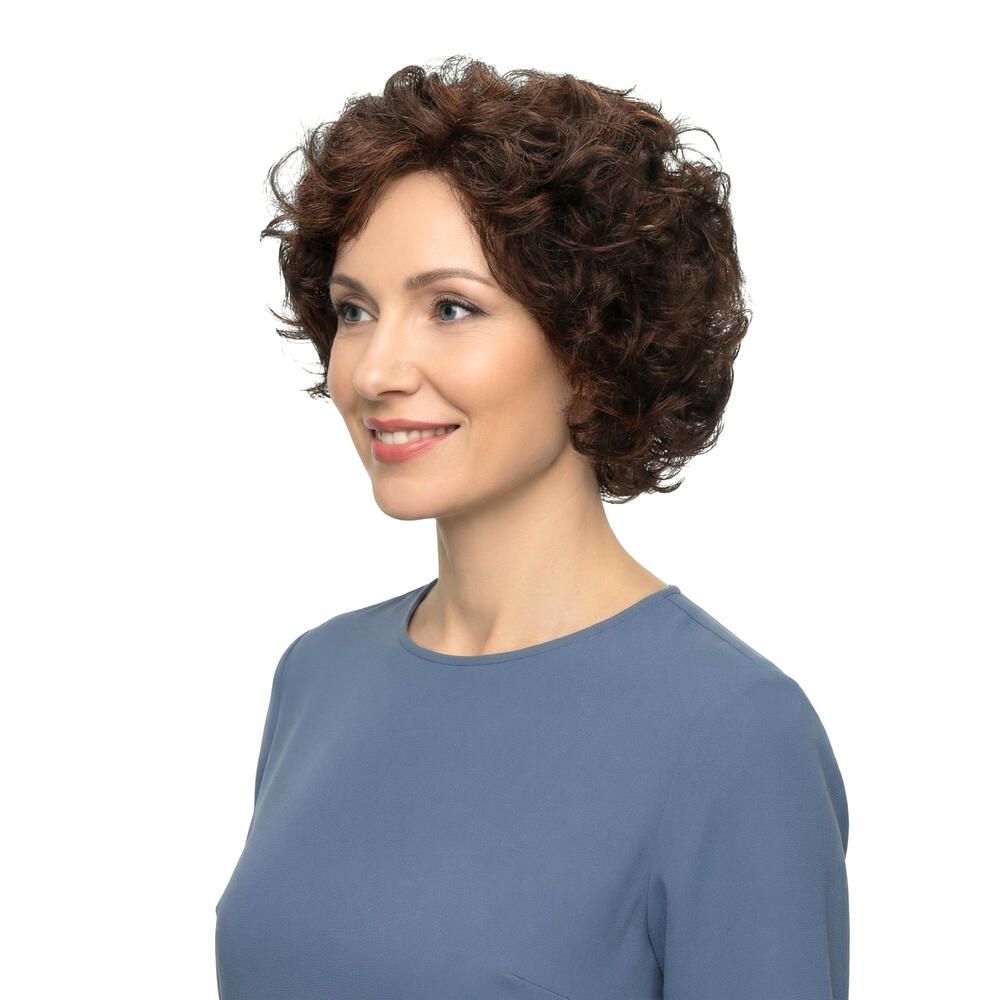 Frau mit brauner, lockiger Perücke. Seitenansicht. Sie trägt ein blaues Oberteil. Sie lächelt. Hintergrund weiß.