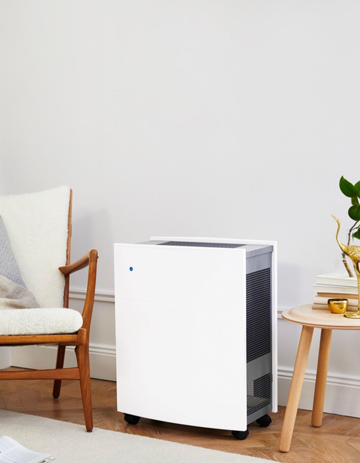 Luftreiniger in Raum. Weißes Gehäuse, Rollen. Steht neben Tisch und Stuhl. Schwarzes Gitter sichtbar.