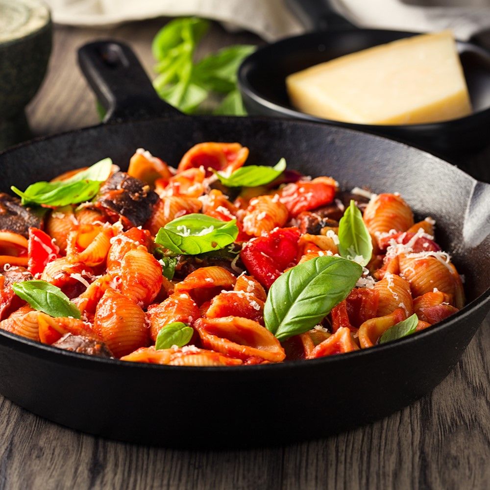 Pasta Gericht in schwarzer Pfanne. Muschelnudeln mit Tomaten, Auberginen und Basilikum. Käse und Basilikumblätter.