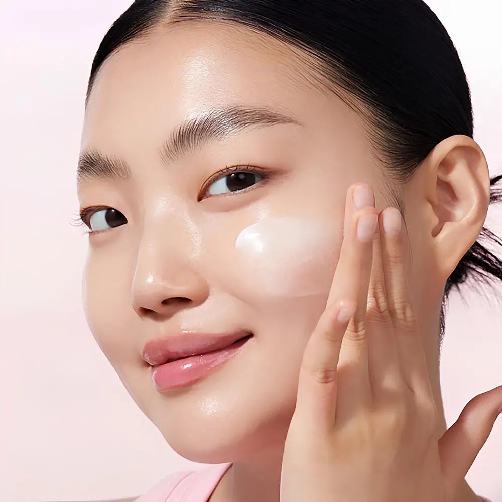 Frau trägt Creme auf ihr Gesicht auf. Rosa Oberteil. Gesicht glänzt. Creme-Anwendung.