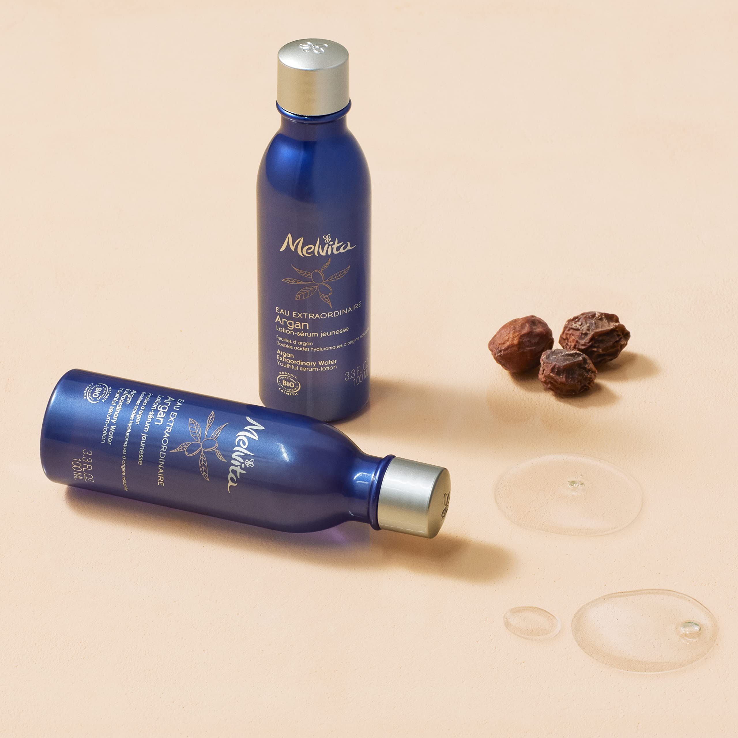 Zwei blaue Flaschen mit silbernen Deckeln. Daneben Argan-Samen und Flüssigkeitstropfen. Aufschrift Melvita.