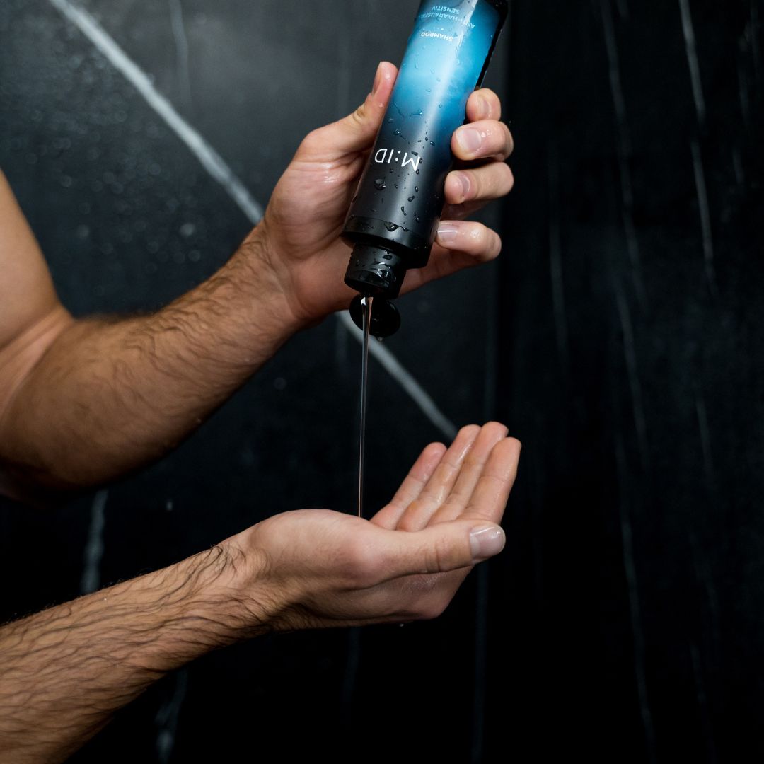 Hände halten eine schwarze Flasche. Produkt wird in die Handfläche gegossen. Aufschrift: M:ID, Anti-Haarausfall Sensitiv Shampoo.